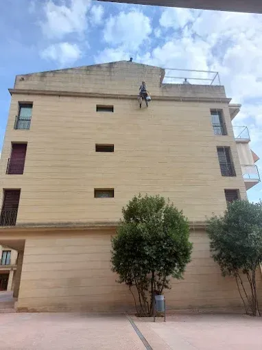 Un trabajador subido a una escalera limpia la fachada de un edificio de apartamentos de color beige bajo un cielo nublado.
