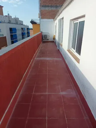 Estrecha terraza en la azotea con suelo de baldosas rojas, paredes blancas y una unidad de aire acondicionado a lo lejos.
