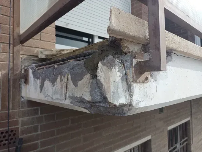 Esquina de balcón de hormigón dañada con barras de refuerzo expuestas y soporte desmoronándose sobre una pared de ladrillo.