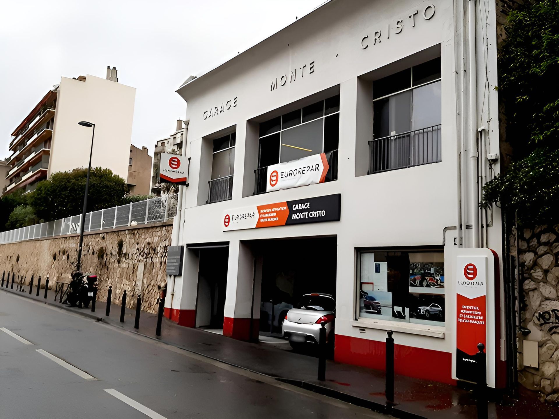 Extérieur d'un bâtiment de deux étages, blanc et rouge. Une voiture est garée au rez-de-chaussée. Des panneaux avec des logos sont visibles.