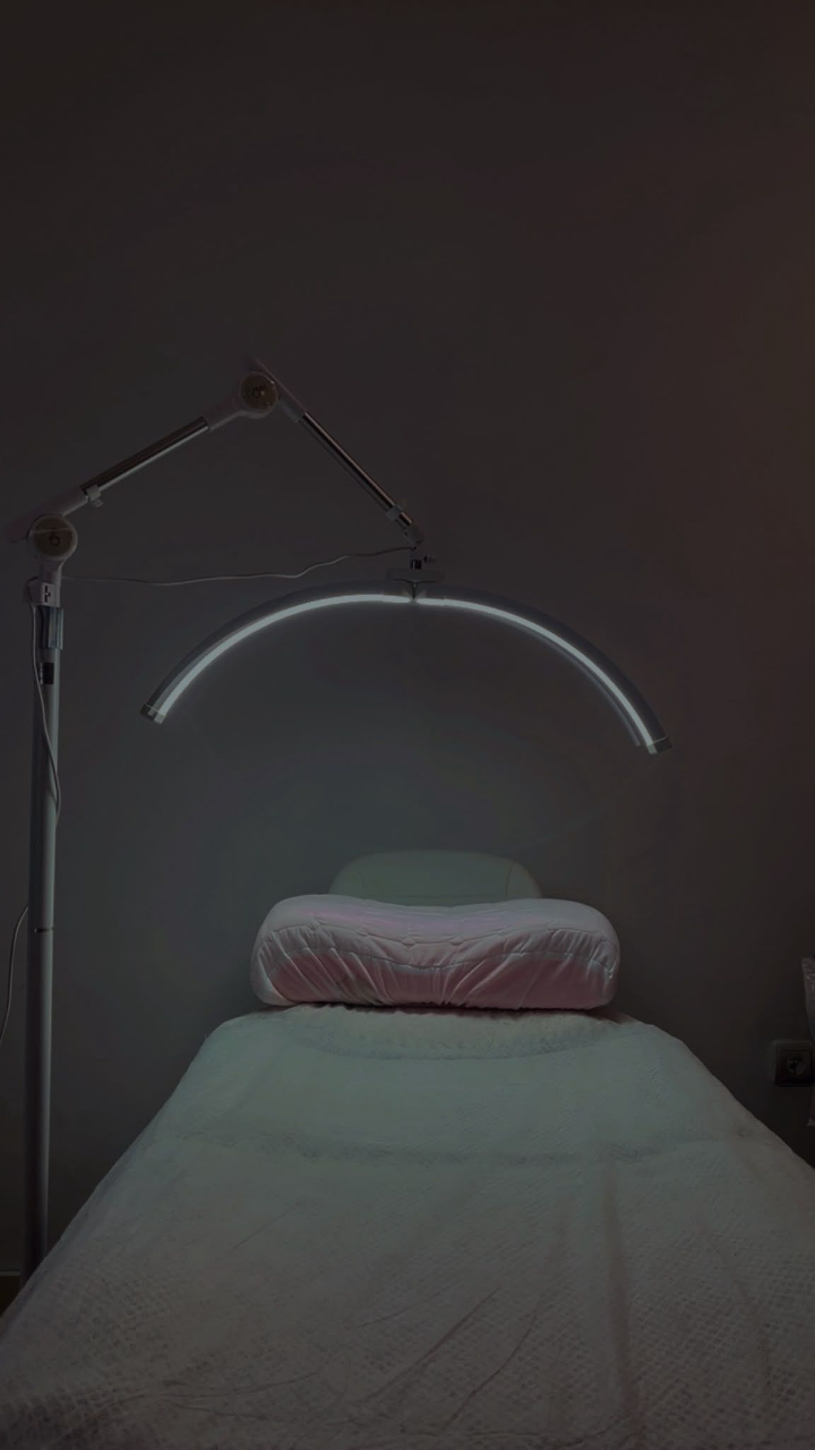Habitación con poca luz, cama hecha y una lámpara curva que emite una luz tenue sobre el cabecero.