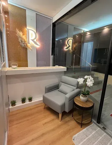Vestíbulo moderno con escritorio blanco, silla gris, suelo de madera y un monograma luminoso en la pared.