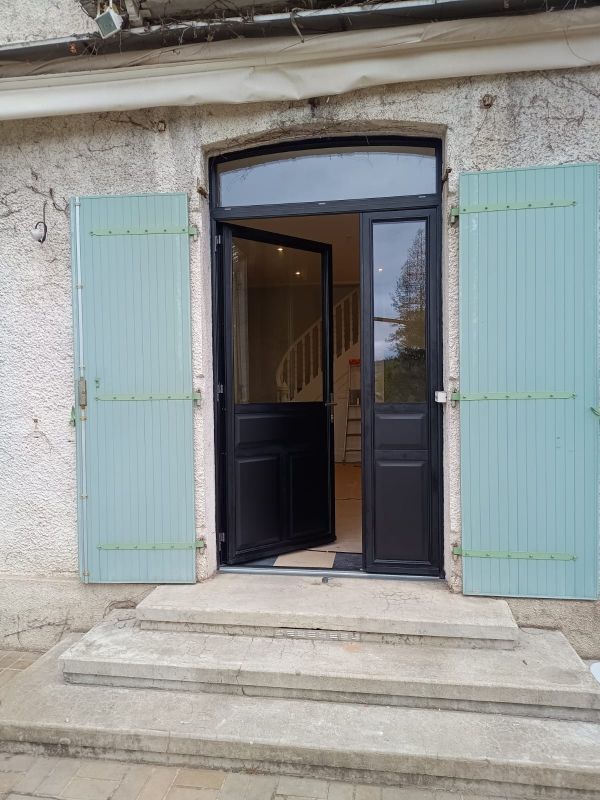 Vue extérieure d'une maison avec une porte noire ouverte, des volets bleus et des marches en pierre menant à l'étage.