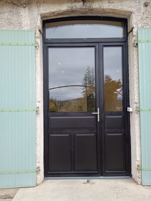 Porte d'entrée noire avec imposte latérale et fenêtre cintrée au-dessus, encastrée dans un mur de pierre et encadrée de volets verts.