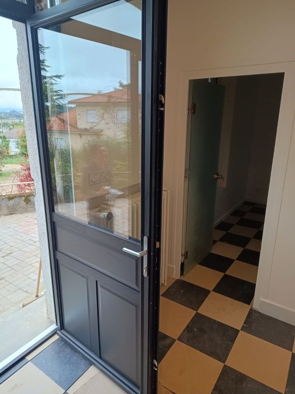 Porte noire et grise, panneaux de verre, donnant sur un couloir au sol à damier, baigné de lumière naturelle.