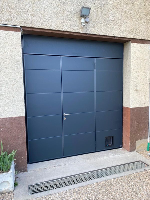 Porte de garage métallique bleue encastrée dans un mur en stuc, avec une petite grille d'aération et d'évacuation en dessous.