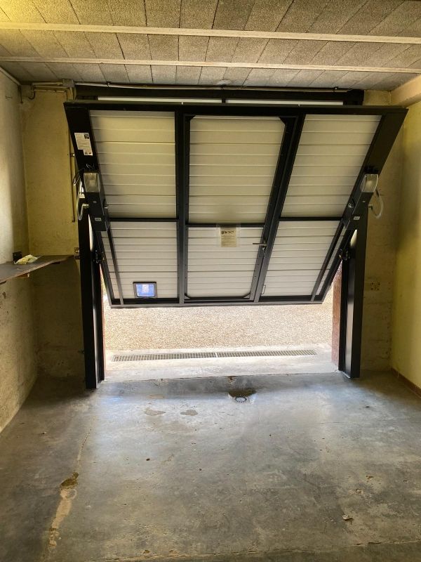 Porte de garage inclinée donnant sur un emplacement de stationnement sombre au sol en béton, éclairé par la lumière extérieure.