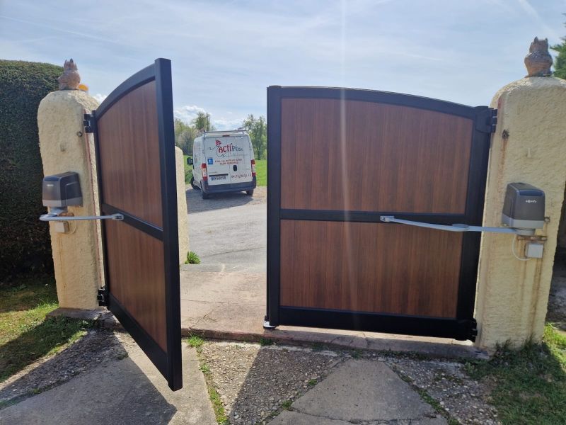 Portail d'allée en bois ouvert entre des piliers de pierre, avec une camionnette visible au-delà.