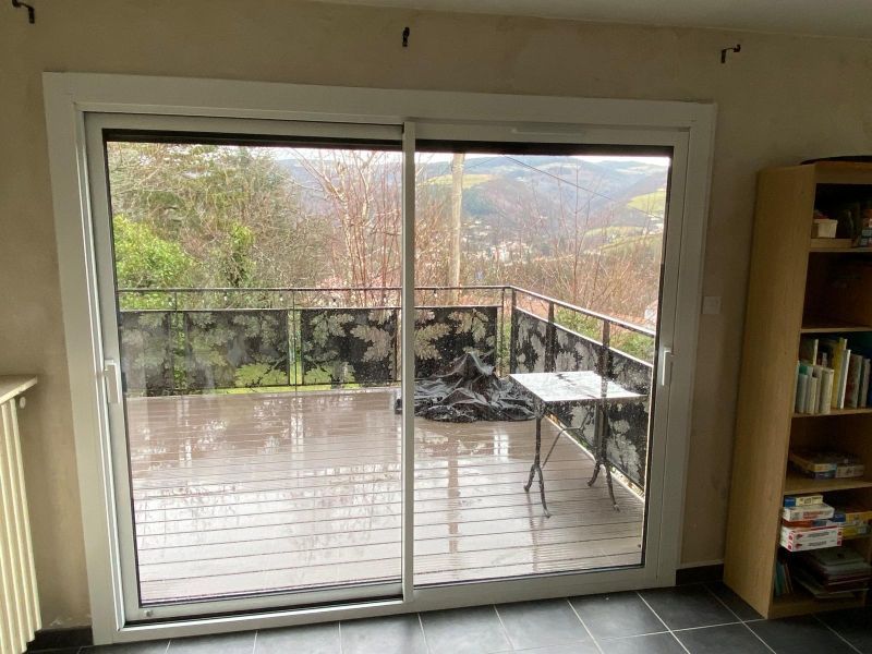 Des portes coulissantes en verre s'ouvrent sur une terrasse exposée à la pluie, avec une rambarde et des arbres au-delà.
