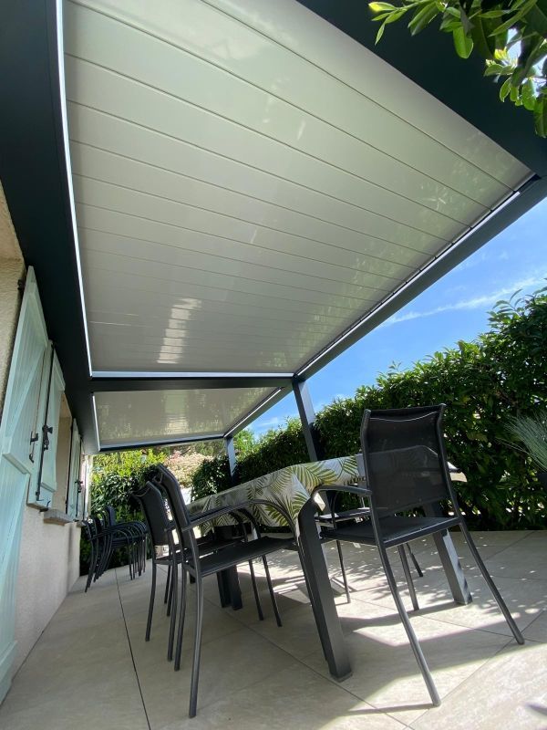 Terrasse couverte avec chaises et table d'extérieur noires, à côté d'une maison, sous une pergola blanche.