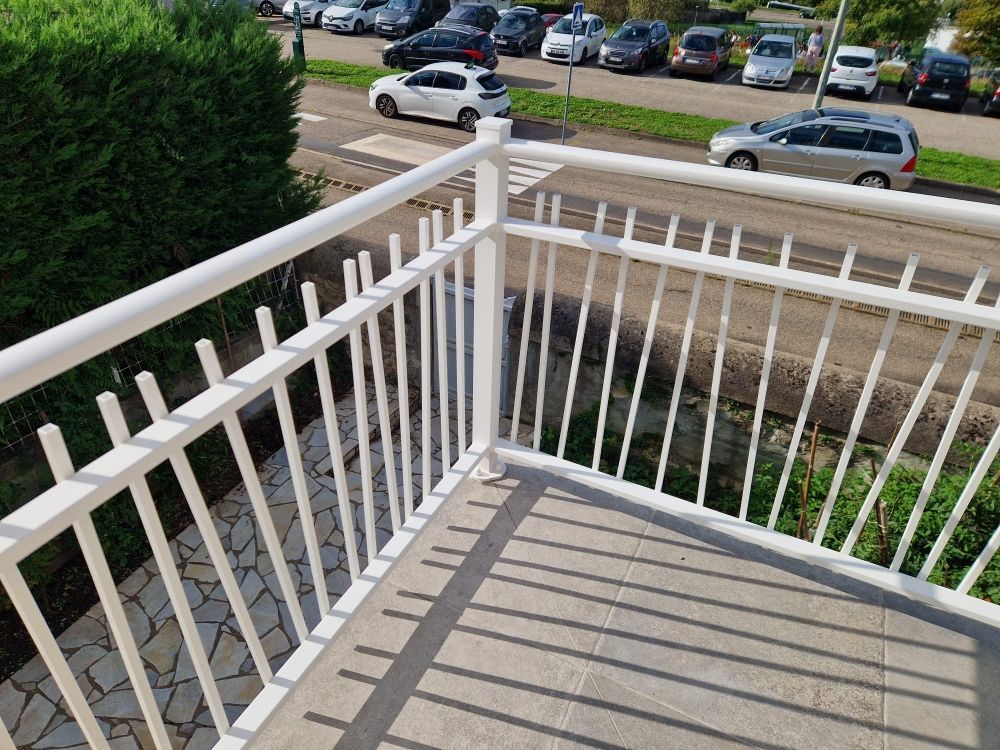 Rambarde de balcon en métal blanc avec sol pavé, donnant sur la rue et les voitures garées.