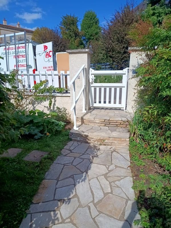 Chemin de pierres menant à un portail blanc muni d'une rampe. Végétation verdoyante environnante. Ciel bleu.