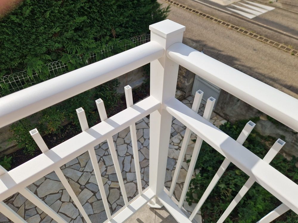 Rambarde de balcon en métal blanc à barreaux verticaux, donnant sur une terrasse en pierre et de la verdure.
