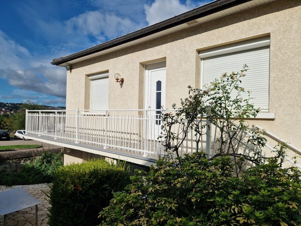 Maison beige avec balcon blanc, porte et volets fermés. Buissons devant.