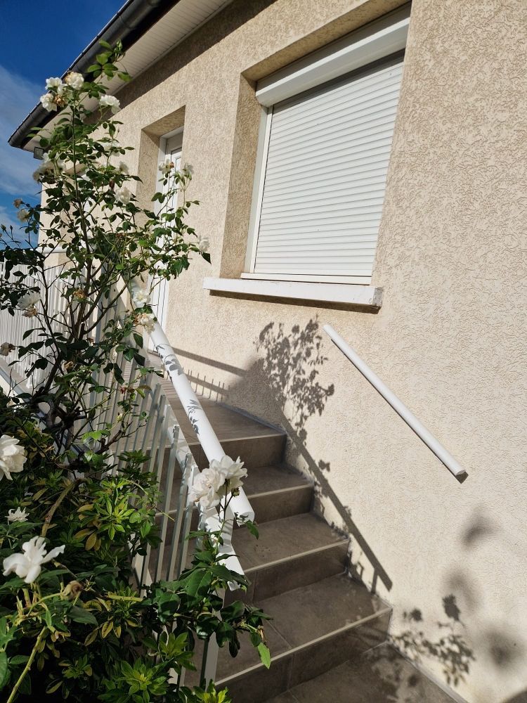 Un escalier extérieur mène à une maison ornée de rosiers blancs et dont le volet est fermé.