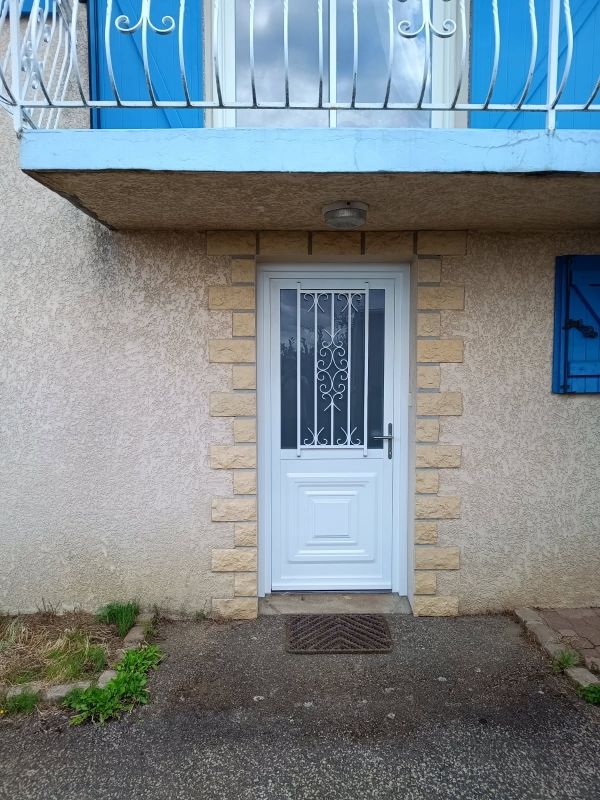 Porte d'entrée blanche avec grille métallique décorative, encadrement en pierre beige, sous un balcon bleu.