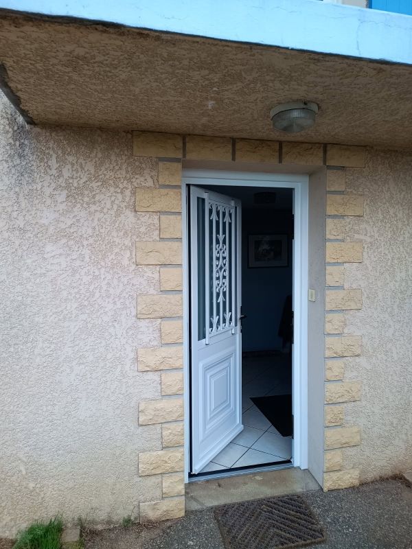 Porte blanche partiellement ouverte avec panneaux métalliques décoratifs, dans un bâtiment extérieur en pierre beige clair et en stuc.