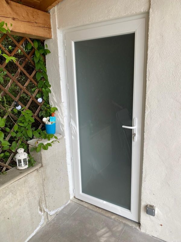 Porte en verre blanc dépoli dans un mur crépi, à côté d'un petit treillis à plantes et d'un coin de patio