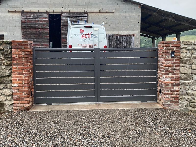 Portail en métal gris entre des piliers en briques devant un bâtiment rustique avec un sac de chantier et une allée en gravier.