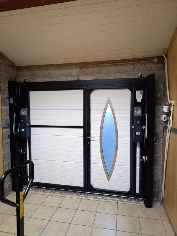 Porte de garage double noire avec un panneau blanc et une fenêtre ovale bleue, installée dans une entrée carrelée.