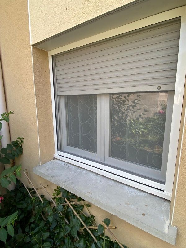 Petite fenêtre avec un volet blanc et un rideau de dentelle, encastrée dans un mur extérieur beige au-dessus d'un rebord en béton.