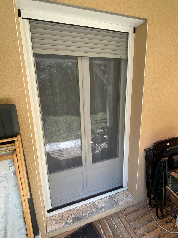 Porte-fenêtre coulissante en verre avec volet gris fermé et encadrement blanc sur un mur extérieur beige