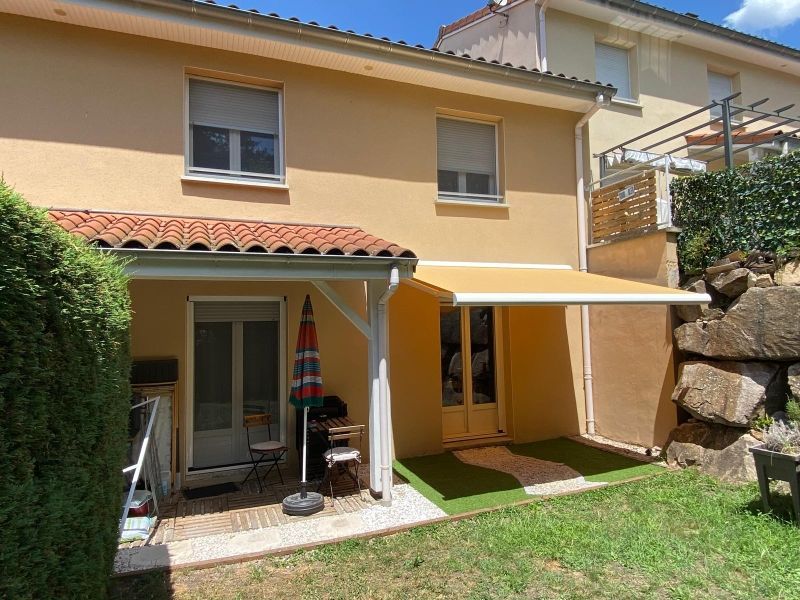 Maison jaune à deux étages avec une terrasse ombragée, un auvent et une petite pelouse dans la cour arrière
