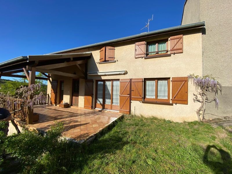Maison rustique baignée de soleil, aux murs beiges, aux volets en bois et appentis couvert donnant sur une cour gazonnée.