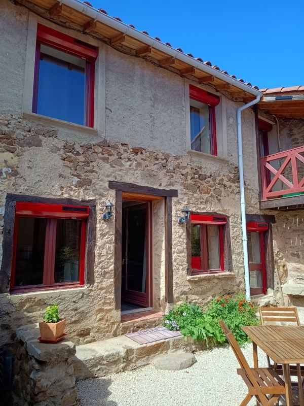 Maison en pierre avec encadrements de fenêtres et volets rouges, porte ouverte et terrasse.