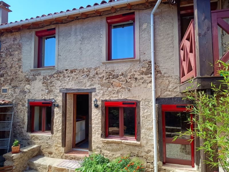 Maison en pierre aux encadrements de fenêtres et volets rouges. Porte ouverte, balcon et plantes vertes.