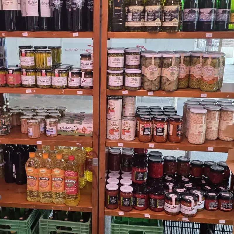 Una estantería de madera en una tienda, repleta de tarros de conservas, botellas de vino y aceite.