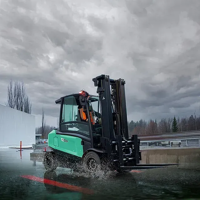 Una carretilla elevadora verde circula por una zona inundada en un día nublado.