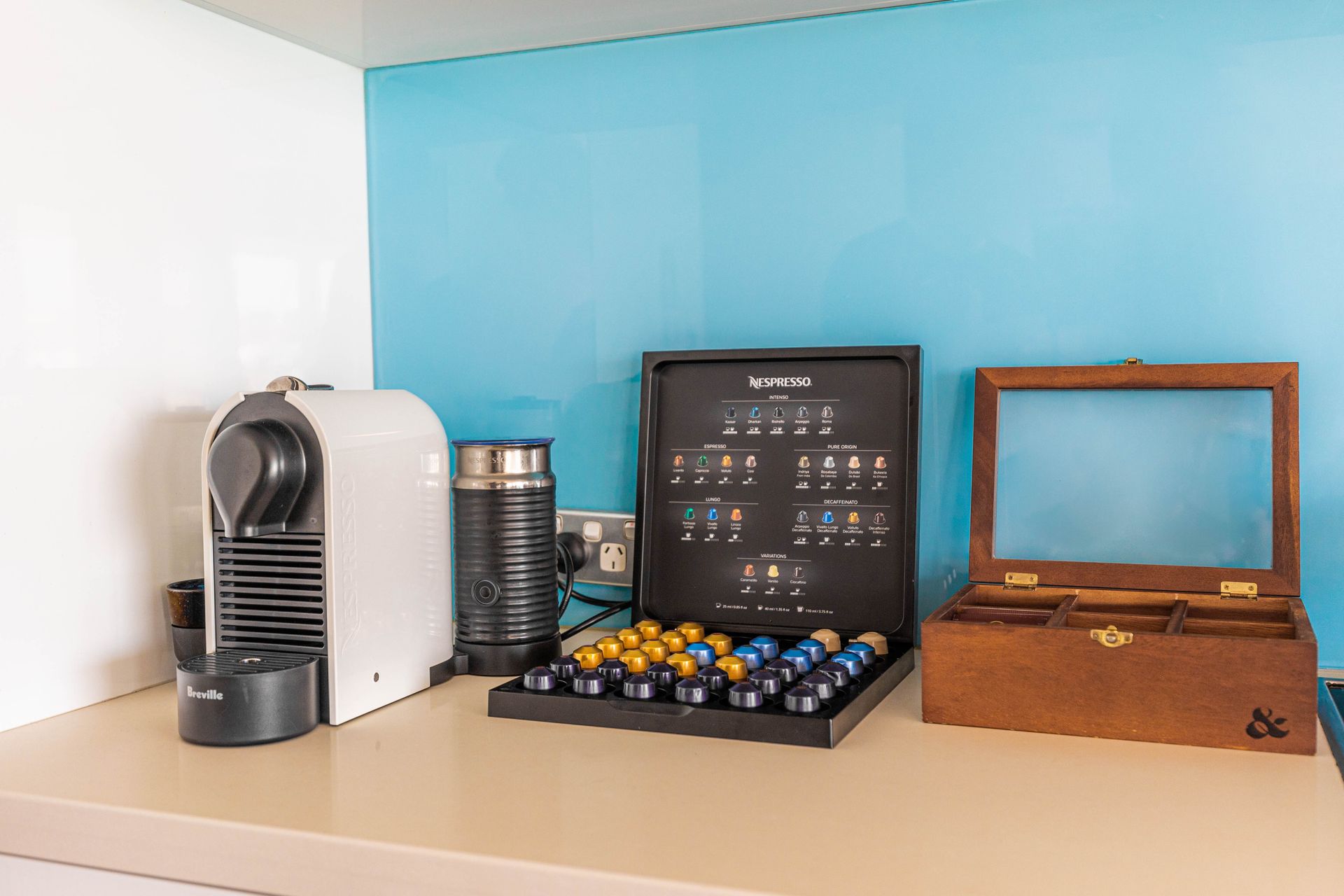 Coffee machine, capsule tray, and wooden box on a shelf against a light blue wall.