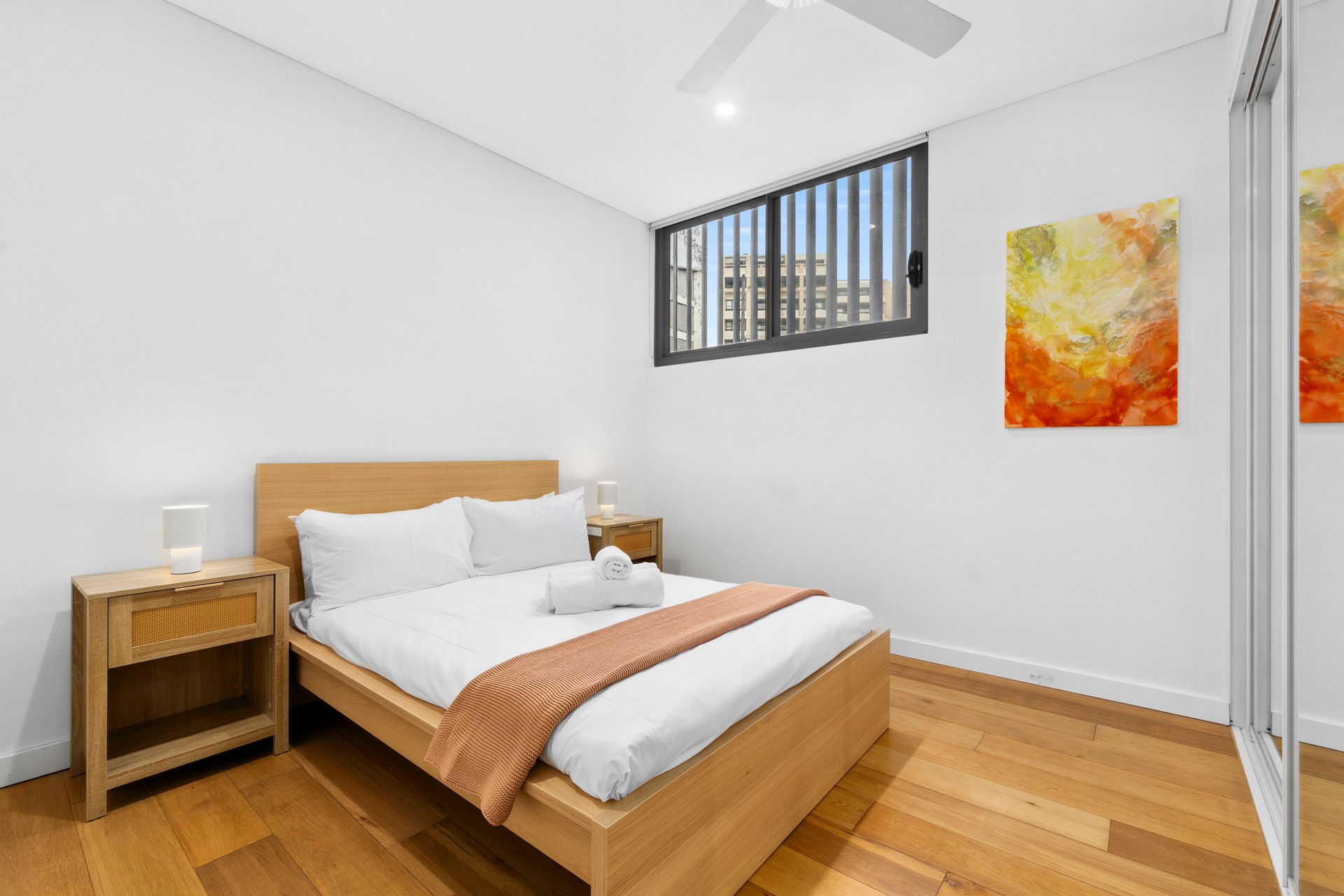Bright bedroom with a wooden bed, nightstand, hardwood floor, and small window with bars.