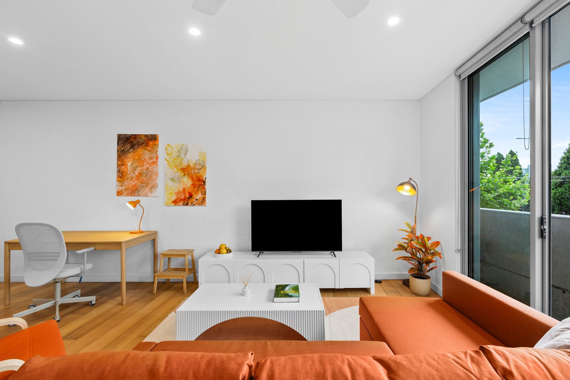 Modern living room with orange sofa, white TV console, desk, and sliding glass door to greenery