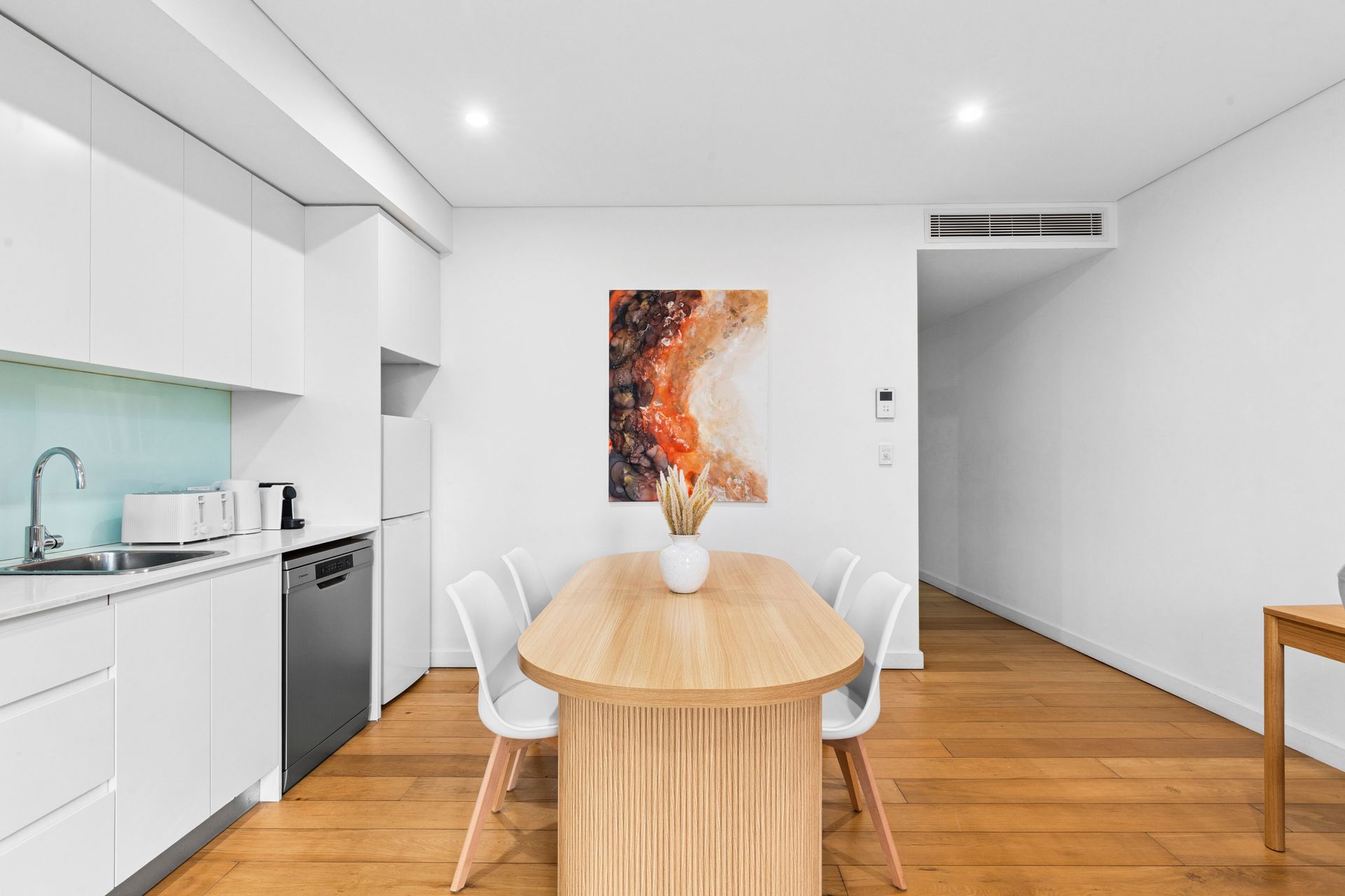 Bright minimalist dining area with wooden table, white chairs, kitchen, and abstract wall art