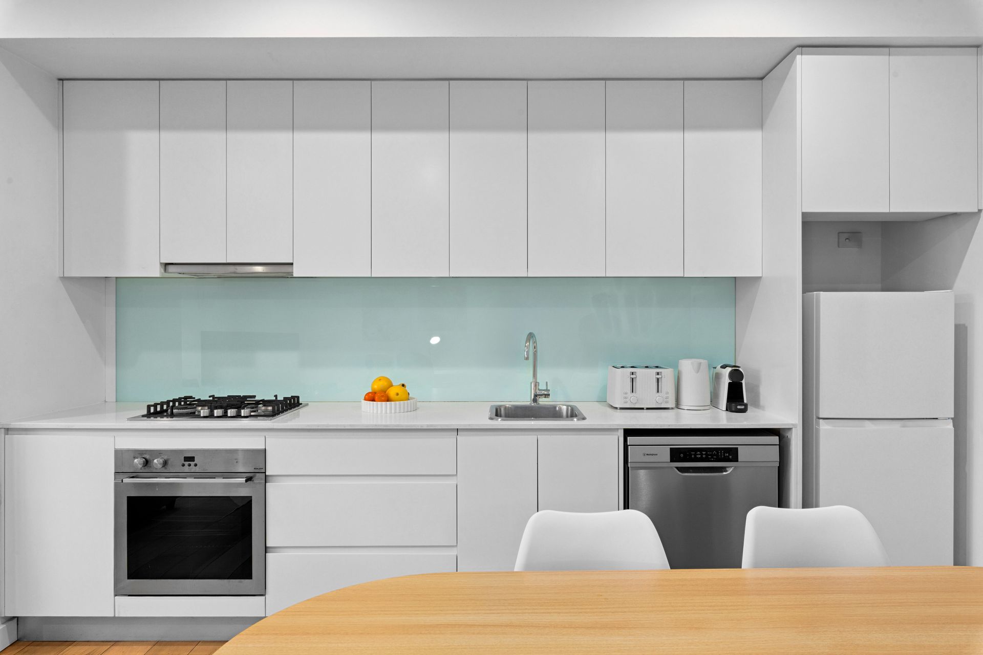Modern white kitchen with wood table, pale green backsplash, and stainless steel appliances
