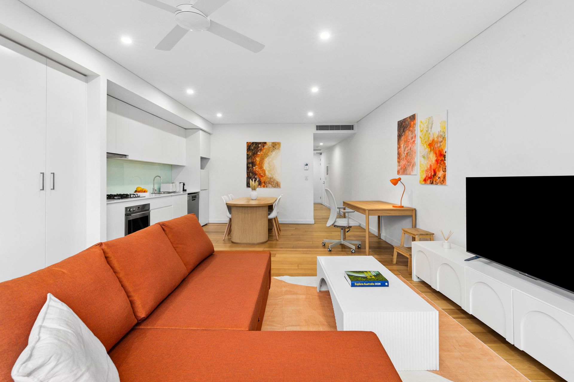 Bright living room with orange sofa, white TV console, dining table, and kitchen area