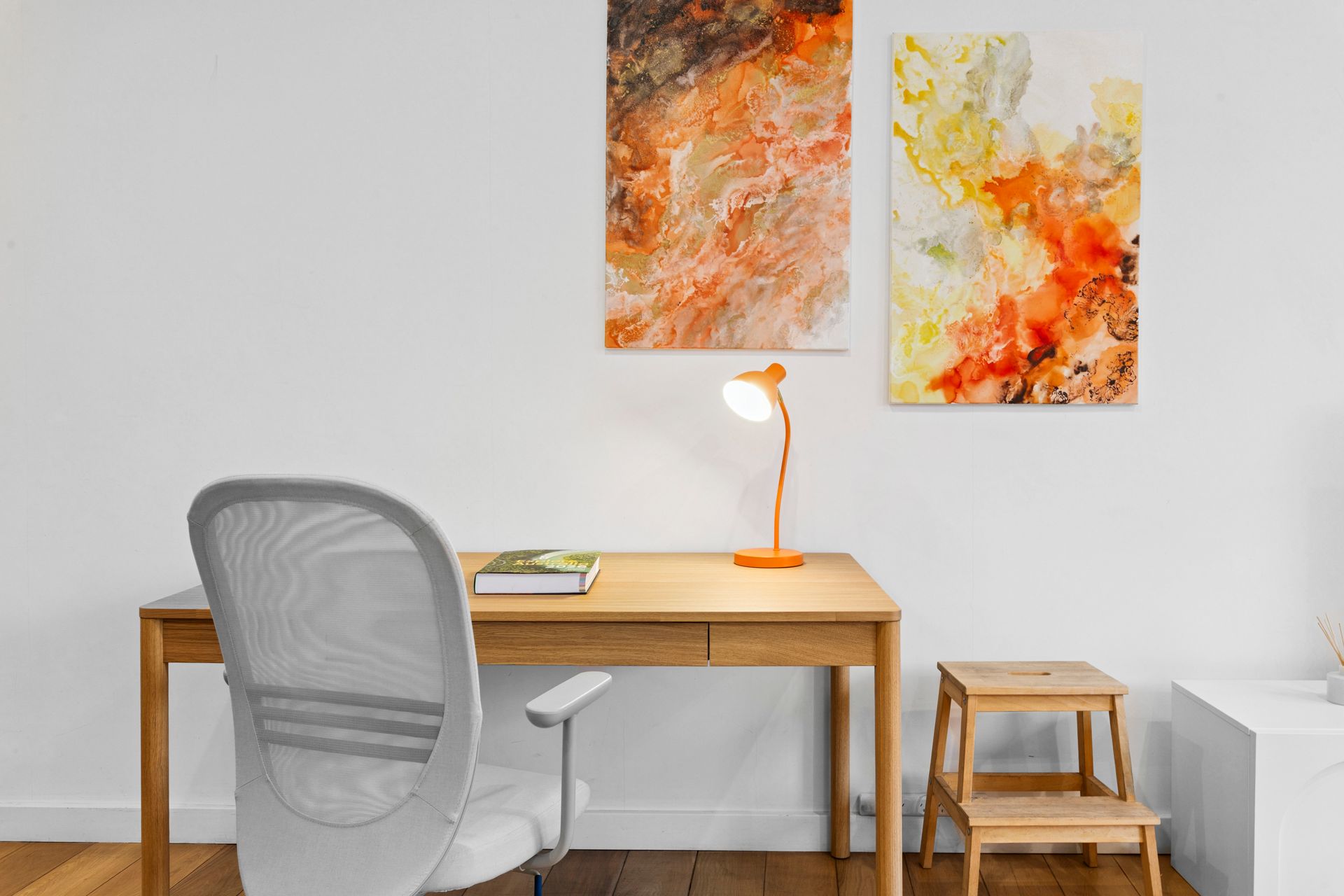 Minimal home office with wooden desk, gray chair, lamp, small stool, and abstract wall art