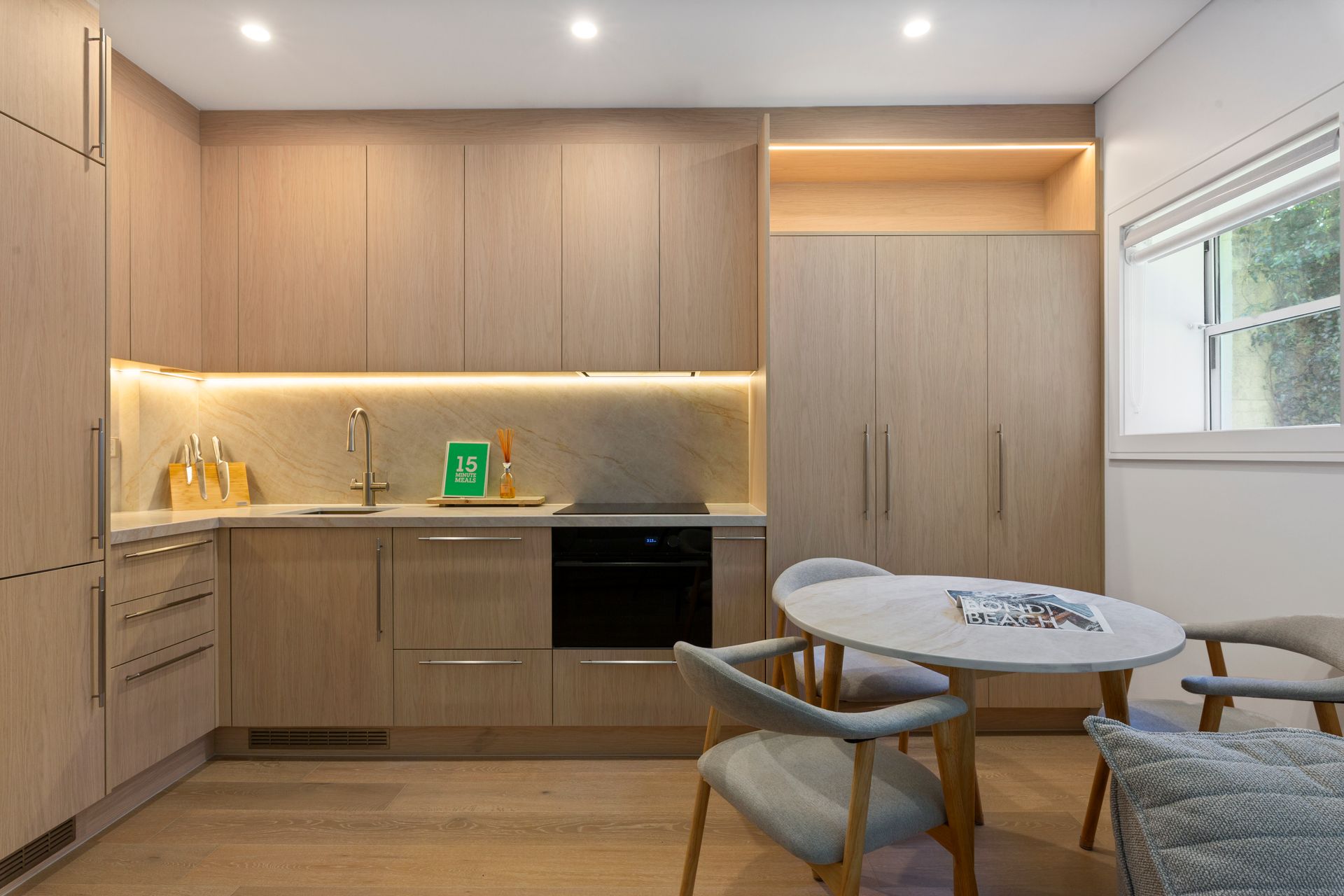 Modern minimalist kitchen with light wood cabinets, black oven, and a round table by a window.