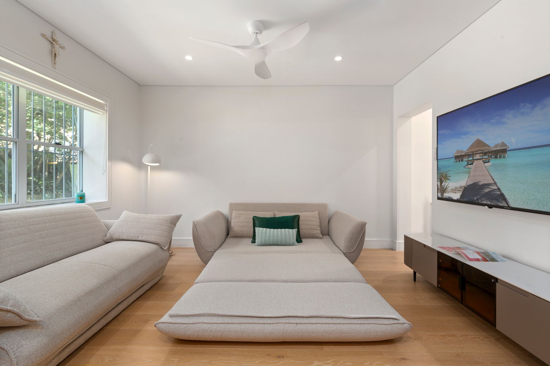 Bright minimalist living room with two gray sofas, a large wall-mounted TV, and a beach scene on screen.
