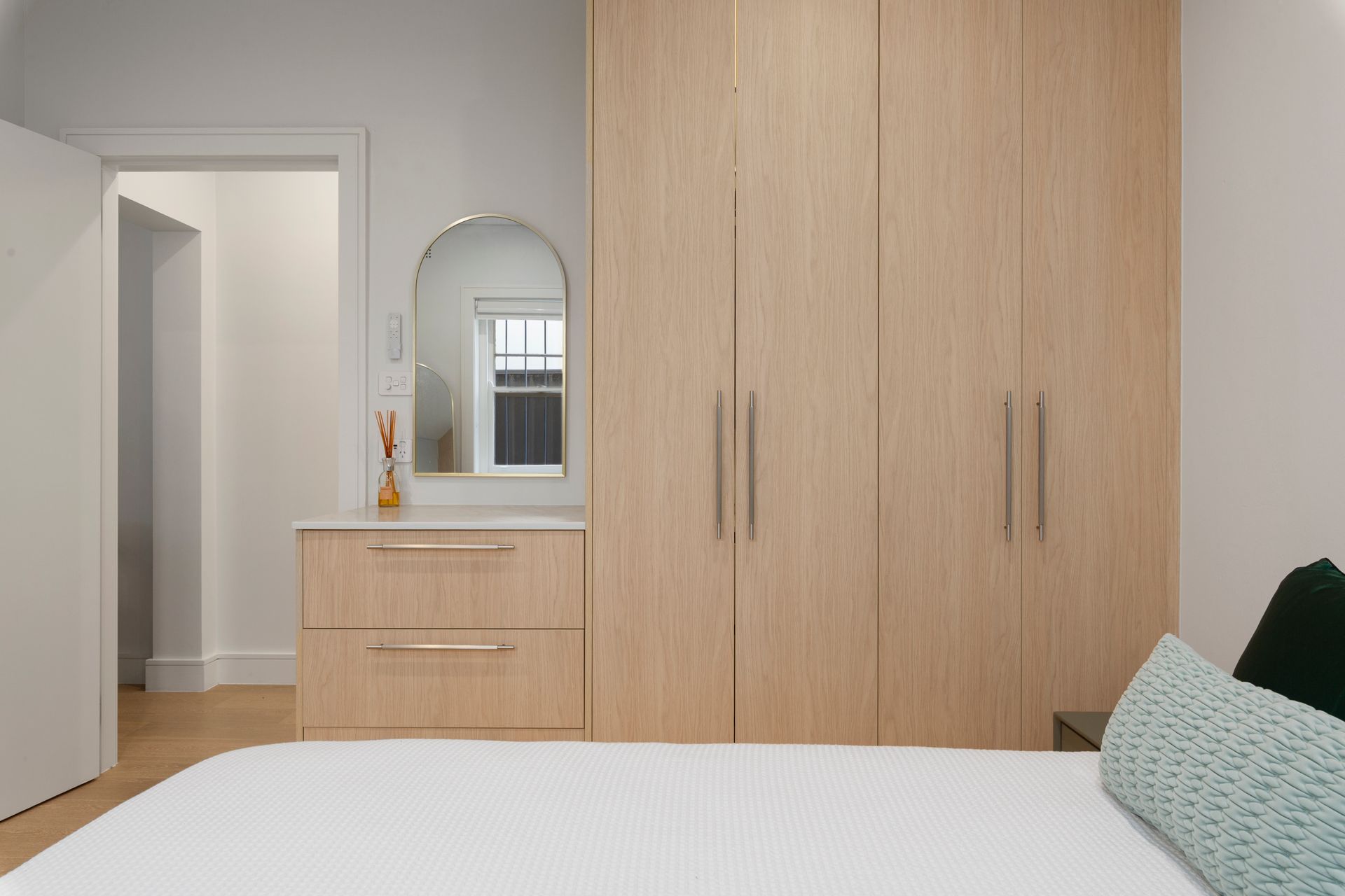 Minimal bedroom with light wood wardrobe, dresser with mirror, and bed in foreground.