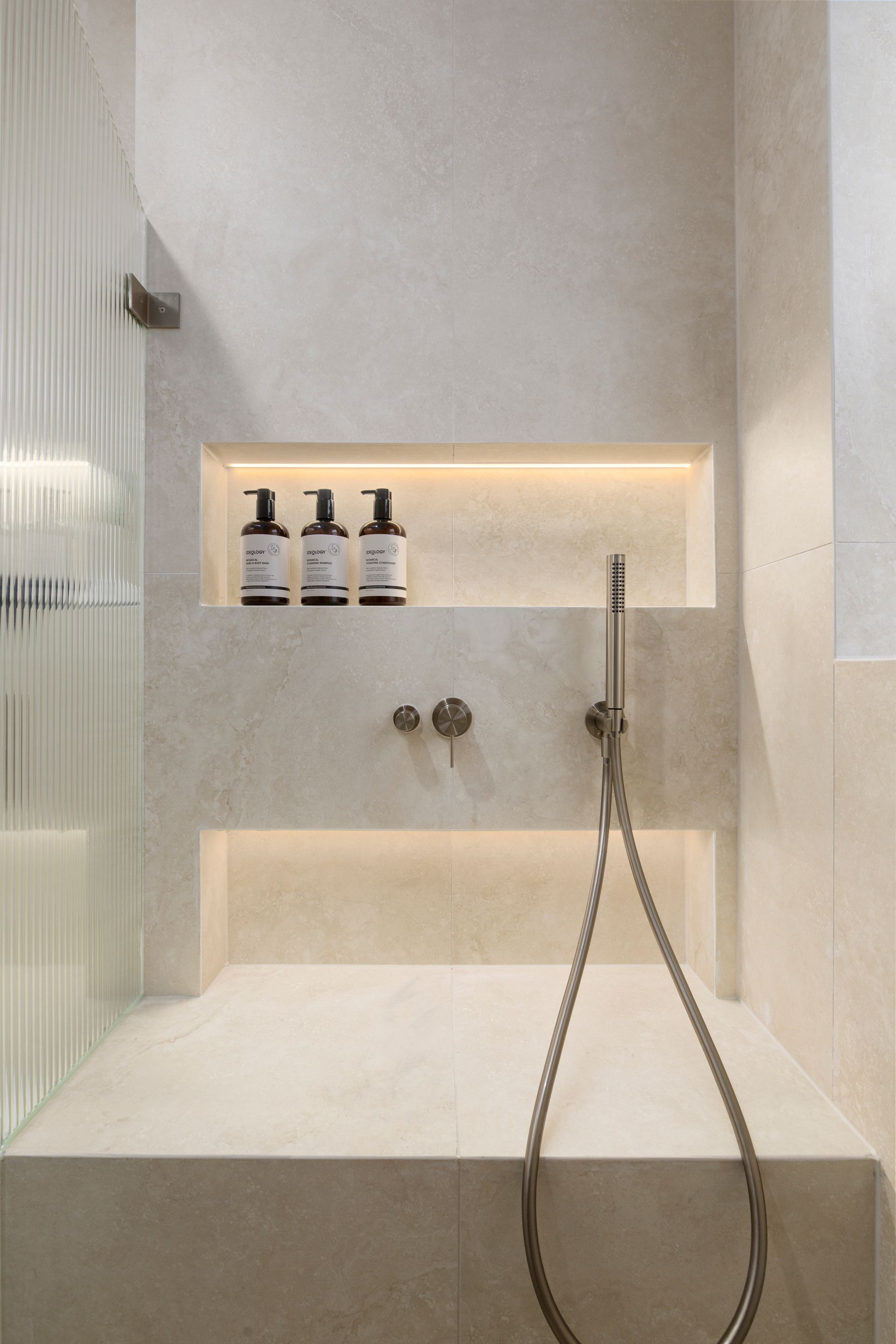 Minimalist white shower with recessed shelf, toiletries, and handheld showerhead.