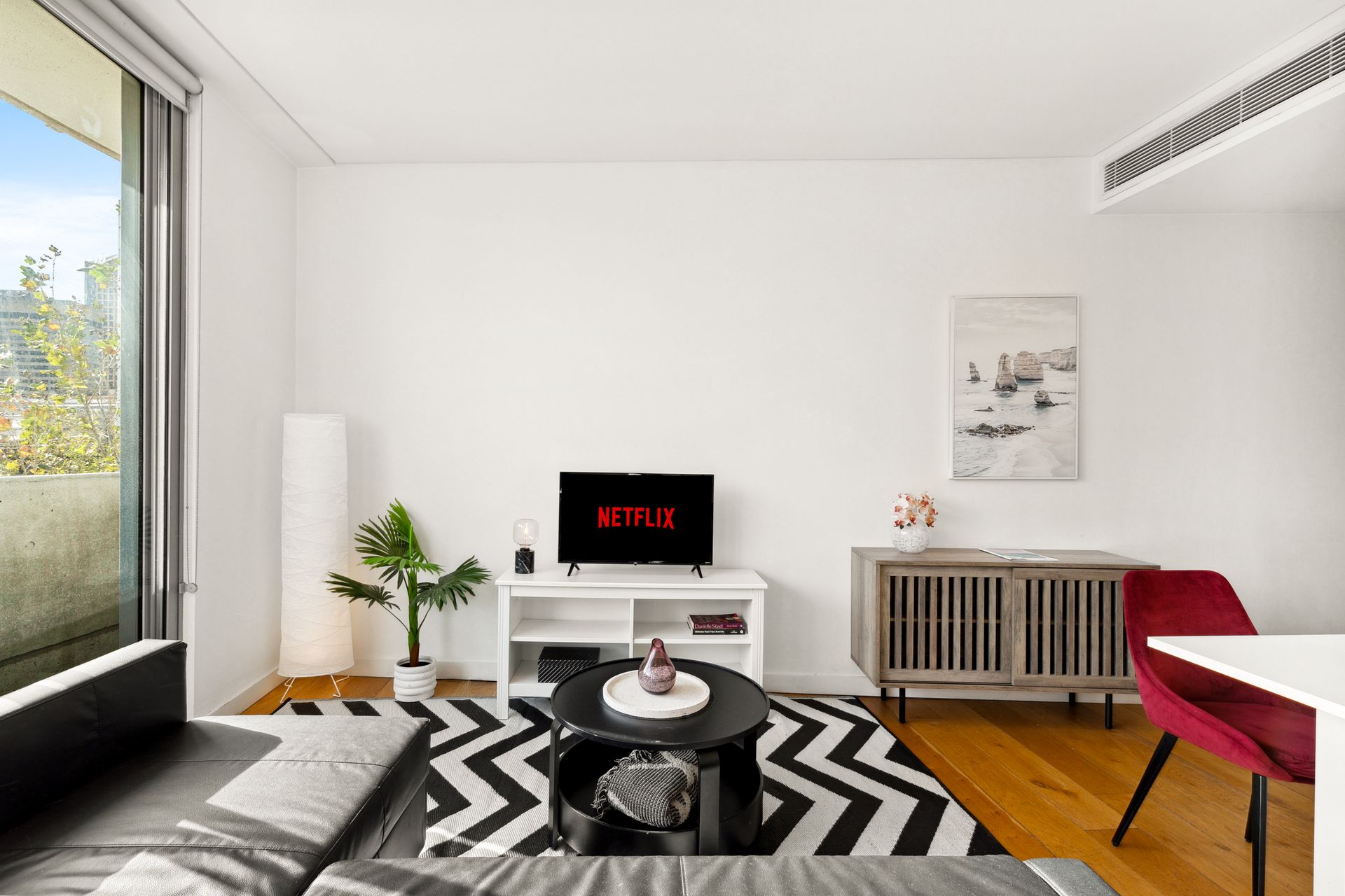 Modern minimalist living room with black-and-white rug, white walls, red chair, and large window.