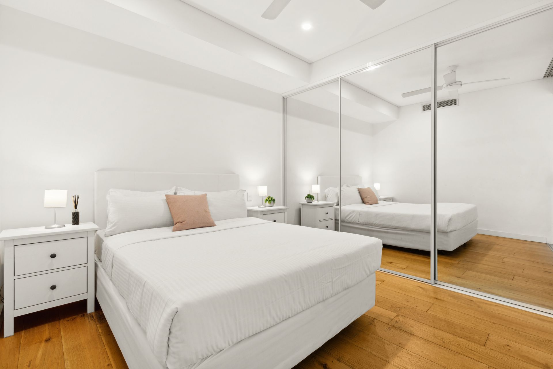 Bright white bedroom with two beds, mirrored closet, bedside lamps, and wood flooring