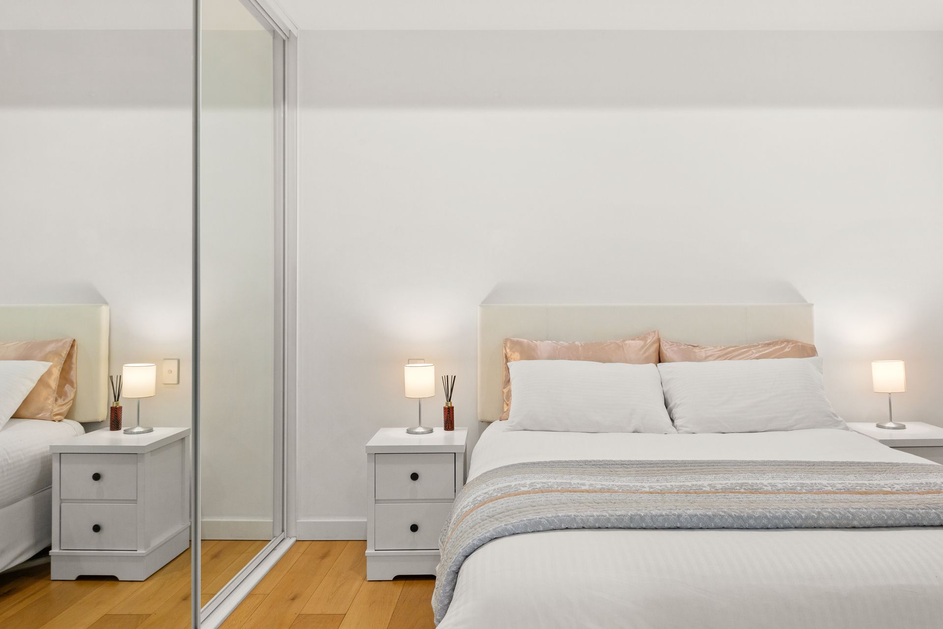 Modern bedroom with a bed, mirrored wardrobe, and two white nightstands with lamps