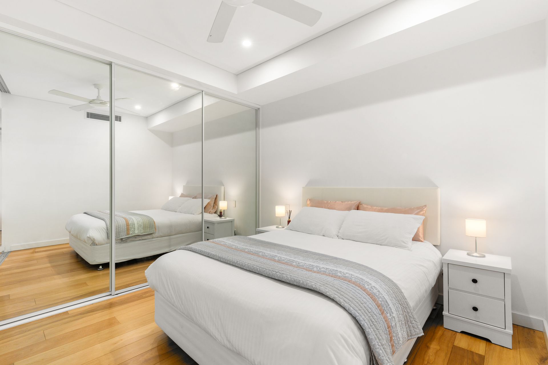 Bright modern bedroom with a bed, mirrored closet doors, and white nightstands on wooden floors