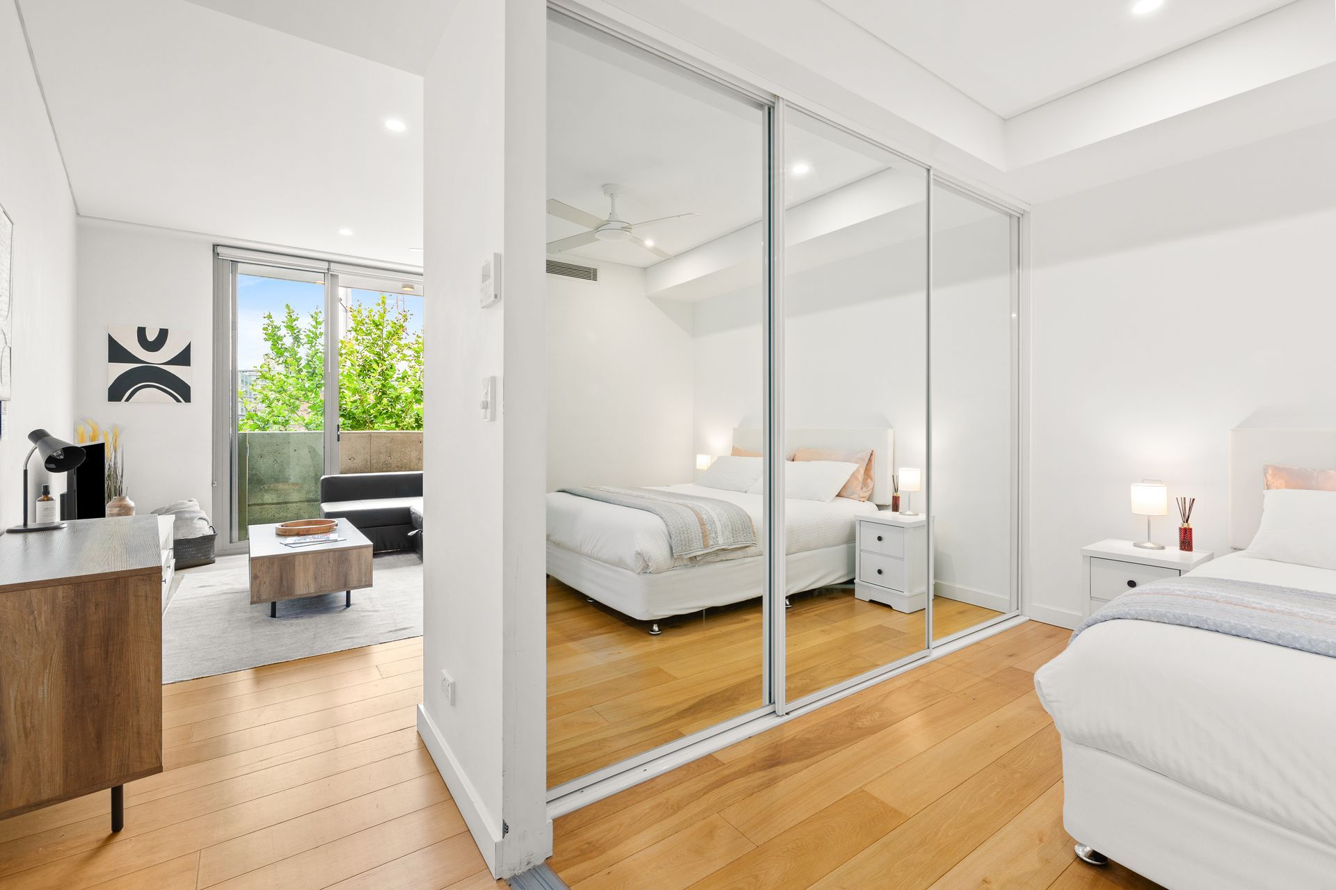 Bright modern bedroom with white walls, mirrored closet doors, and wood floors, opening to a seating area and window.