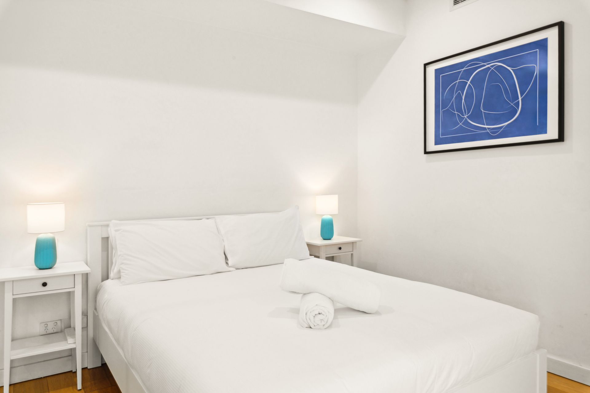 Bright white bedroom with a bed, bedside lamps, and blue wall art