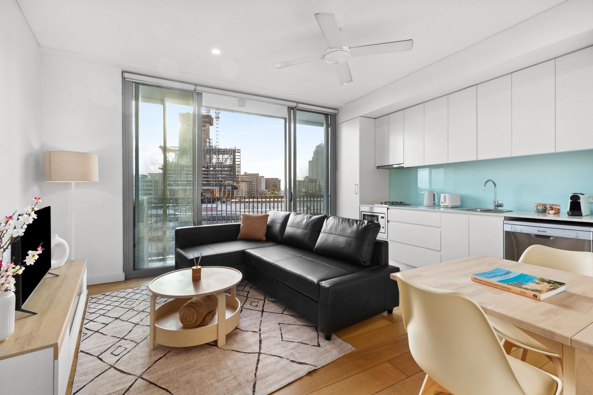 Bright modern living room with black sofa, round coffee table, dining area, and balcony doors overlooking a city skyline